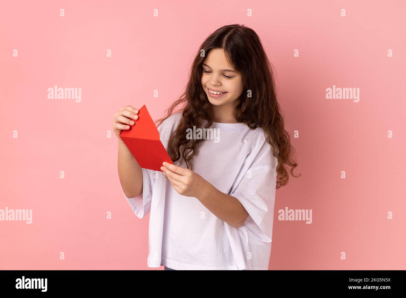Ritratto di bambina che indossa una T-shirt bianca che legge una lettera o un biglietto di auguri, che tiene una busta, sorridendo e gioendo una piacevole notizia. Studio in interni isolato su sfondo rosa. Foto Stock