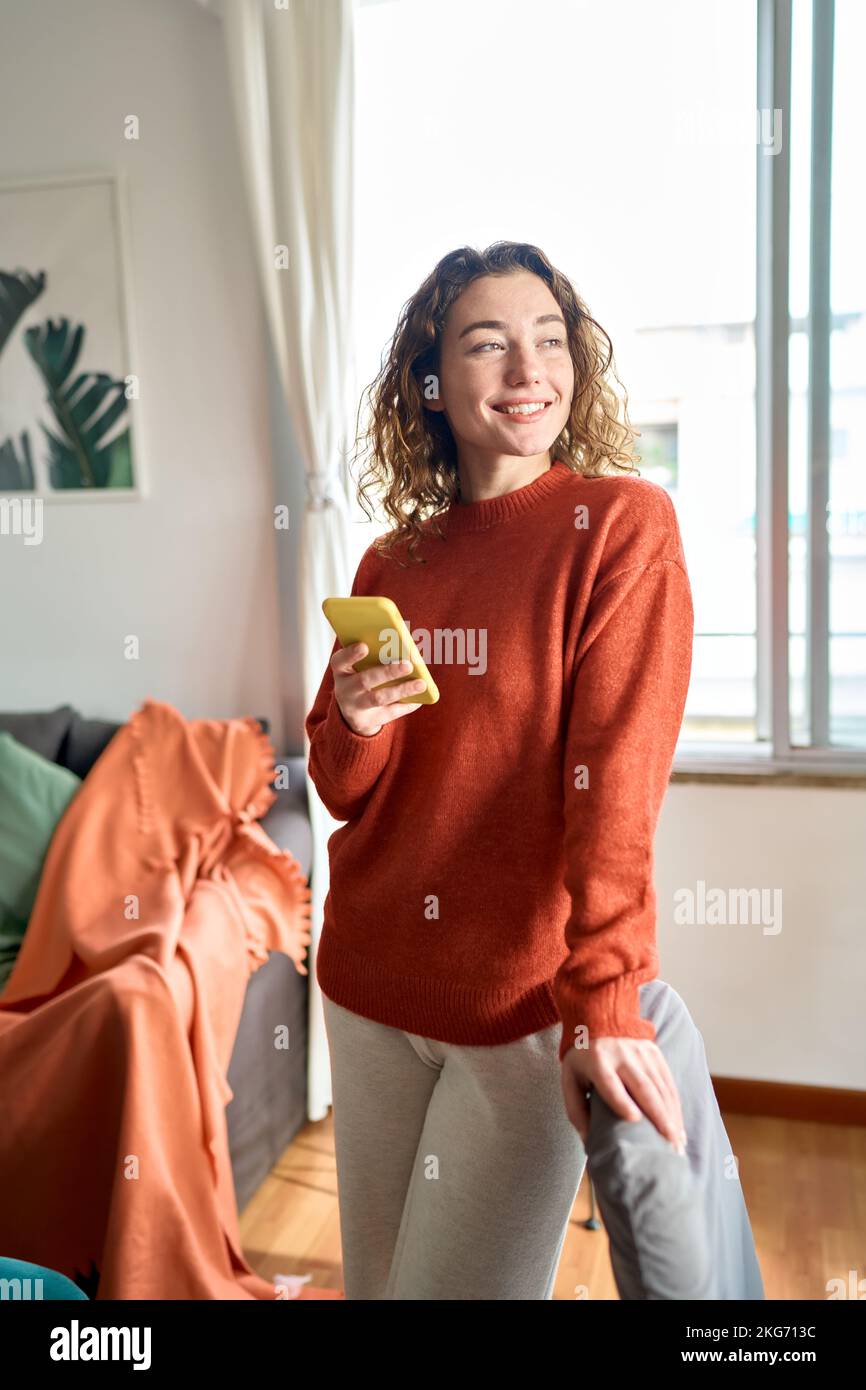 Giovane donna sorridente che usa un telefono cellulare in piedi nel salotto di casa. Foto Stock