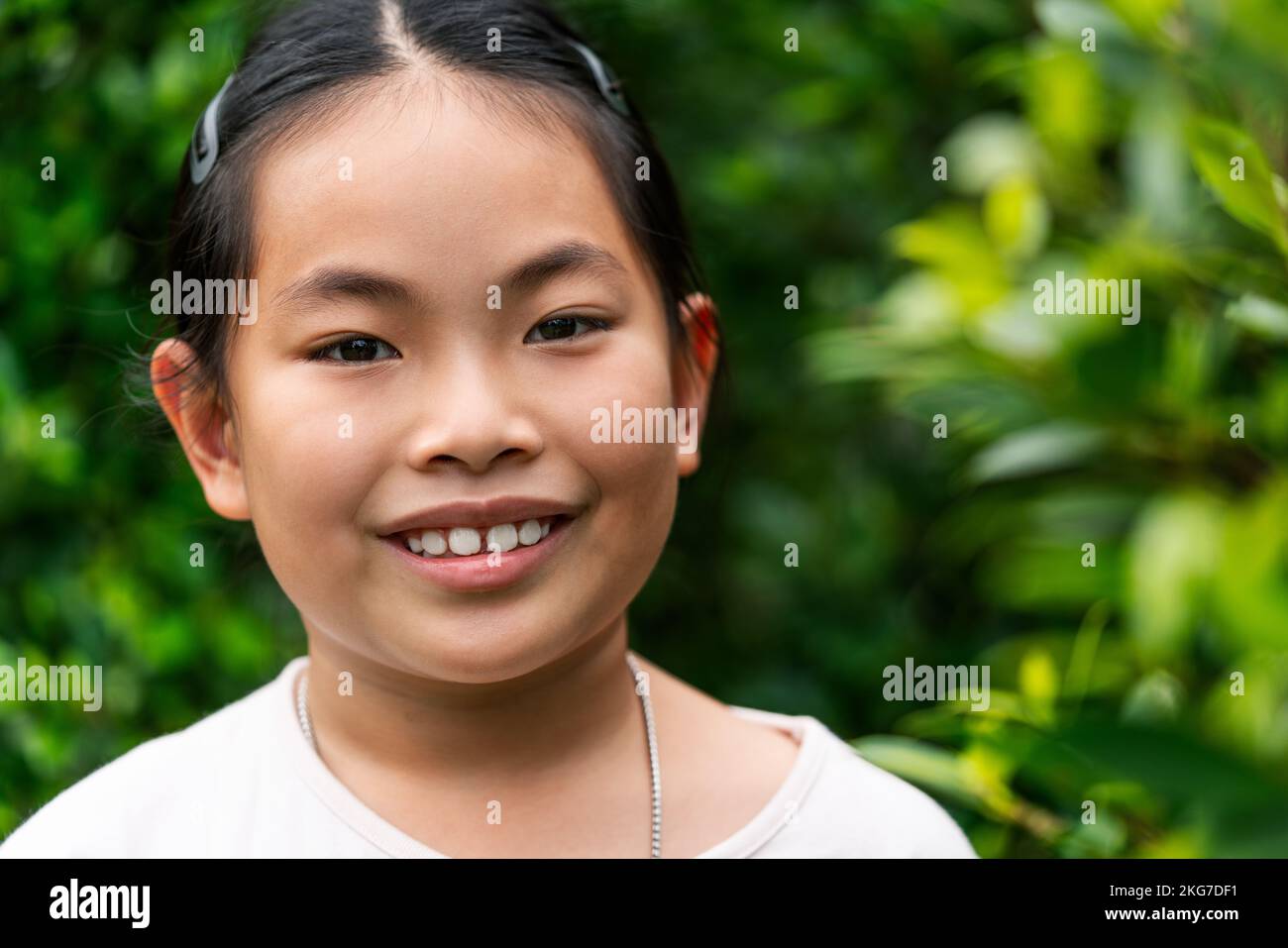 Ritratto di bambina asiatica da 8 a 9 anni, primo piano, sorridente, guardando la macchina fotografica, immagine esterna, sfondo sfocato di verde garde pianta Foto Stock