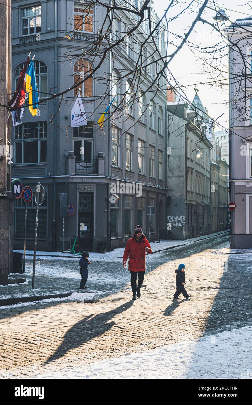 Padre e bambini camminano per strada alla luce del sole nella città vecchia di riga, in Lettonia, in inverno con bandiere, neve e ombre, verticale Foto Stock