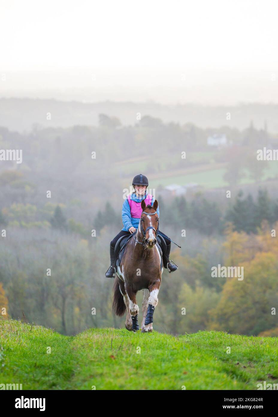 Vista frontale di una donna sorridente cavaliere cavalcata su una riva d'erba con sfondo nebboso Foto Stock