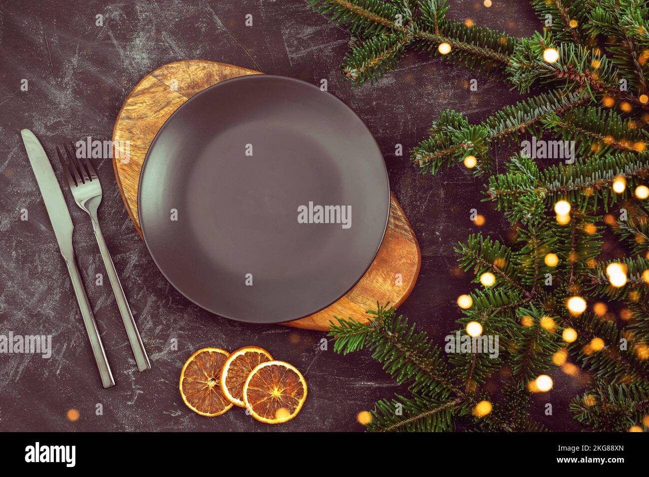 Tavola nera con piatto su tagliere, forchetta e coltello, rami di abete natalizio e fette d'arancia. Vista dall'alto, disposizione piatta Foto Stock