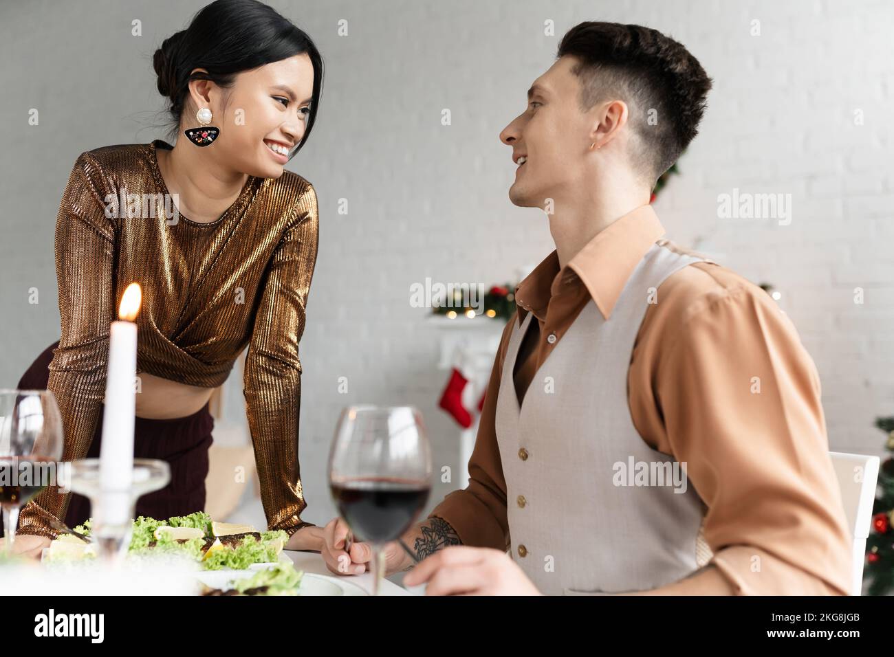 Coppia interrazziale allegra che si guarda l'un l'altro vicino a pasto festivo e bicchieri di vino durante Natale Foto Stock