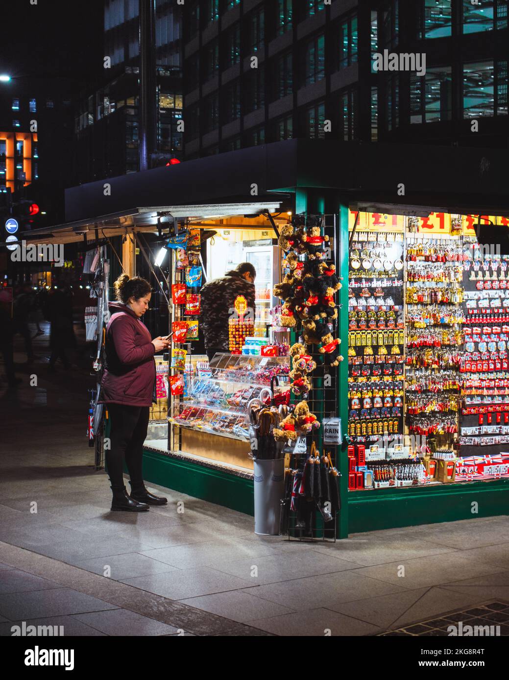 Una donna in un cappotto verde/marrone bruno bruna in un punto vendita della concessione che vende souvenir di notte a Londra. Foto Stock