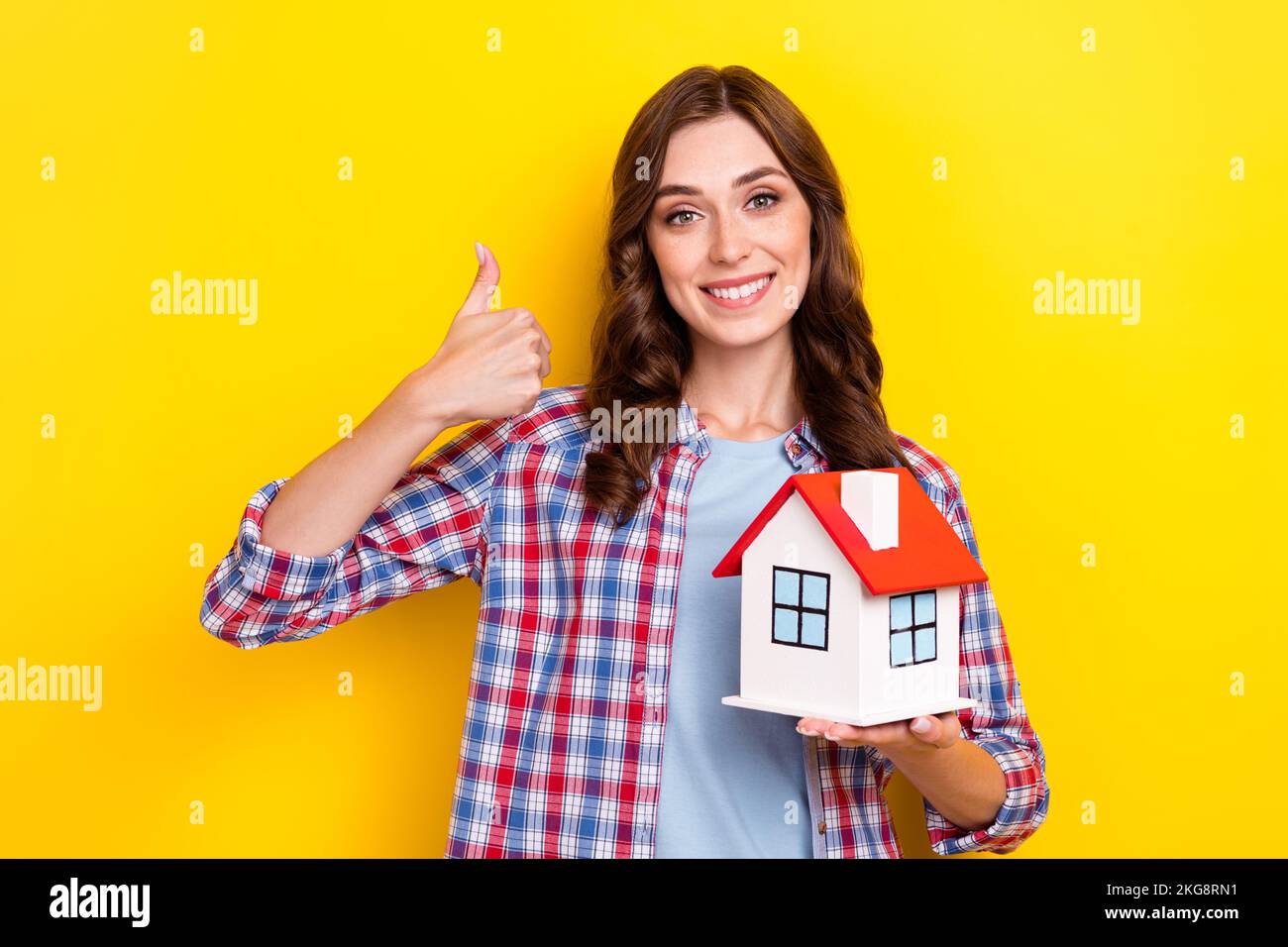 Foto di carino donna indossare camicia a plaid aperto pollice in su tenendo braccia poco rosso tetto casa isolato giallo sfondo colore Foto Stock