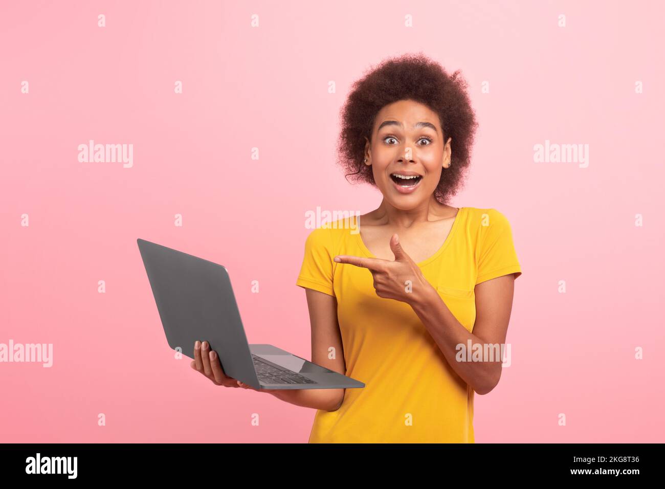 Felice sorpresa giovane afroamericana signora ricciola in t-shirt gialla punta dito sul portatile, gioisce la vittoria Foto Stock