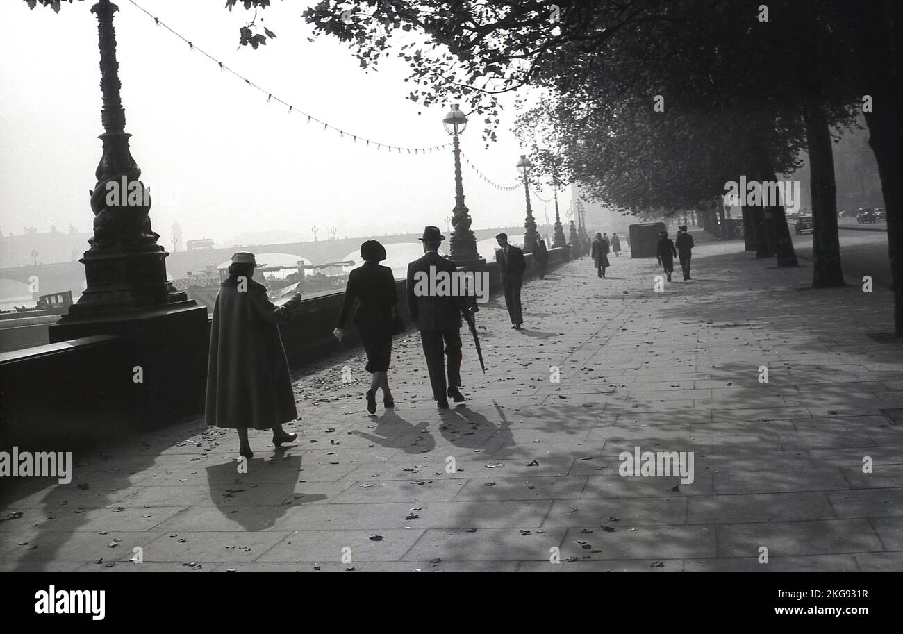 1950s, storico, un Signore e una signora elegantemente vestiti, entrambi indossati ha, camminando lungo l'ampio marciapiede a Victoria Embankment, una passeggiata lungo il fiume Tamigi, Westminster, Londra, Inghilterra UK. Foto Stock