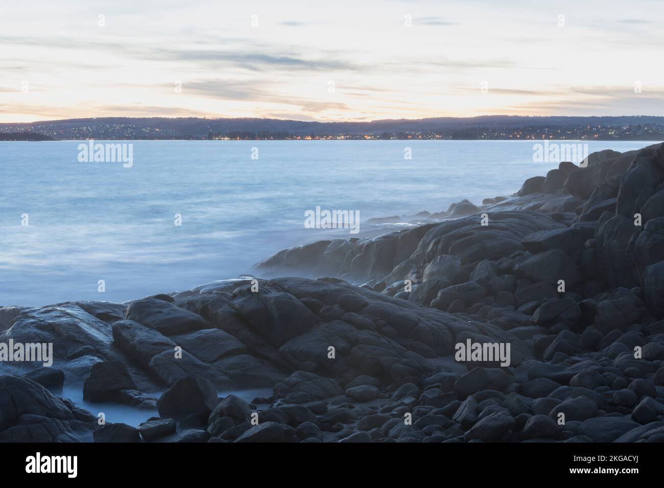 Le onde si rompono sulle rocce di granito a Rocky Bay, Port Elliot, al tramonto guardando verso Victor Harbour e Granite Island, Sud Australia. Foto Stock