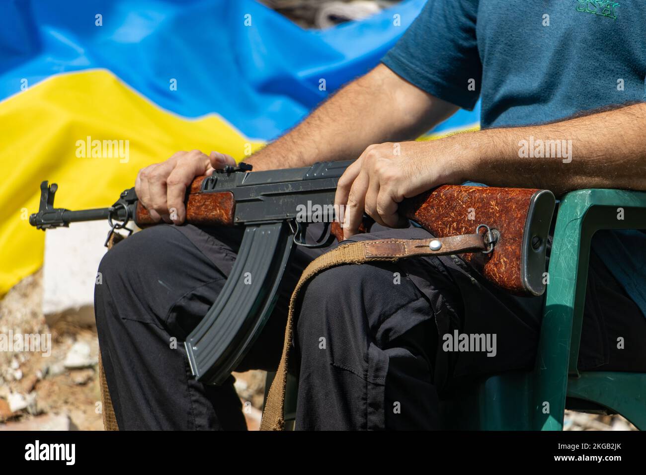 Un uomo con un fucile d'assalto siede di fronte alle rovine con una bandiera Ucraina Foto Stock