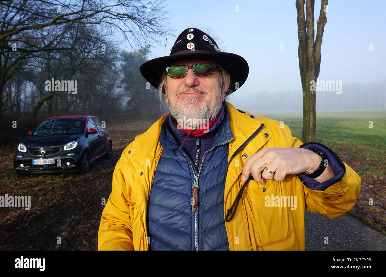 Itterbeck, Germania - Febbraio 28 2021 Un uomo con un impermeabile giallo e un cappello nero da cowboy con spille davanti ad un'auto elettrica nera: Un EQ Smart forfour Foto Stock