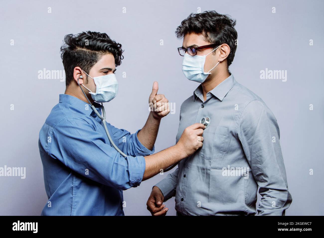 Un cardiologo maschile che indossa una maschera medica esamina il battito cardiaco di un paziente di cuore maschile utilizzando uno stetoscopio e dà un segno di approvazione con un pollice-up g Foto Stock