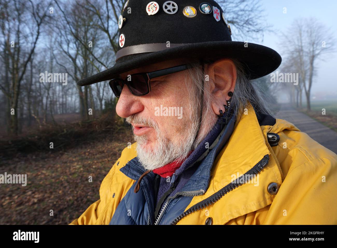 Un uomo anziano con occhiali da sole, un cappello occidentale con spille, orecchini neri e un impermeabile giallo è in piedi in un paesaggio invernale Foto Stock