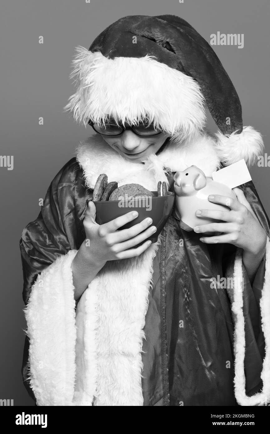 ragazzo giovane e carino babbo natale con bicchieri in maglione rosso e cappello di natale di capodanno con una banca di maiale rosa piggy e biscotti al cioccolato hip in ciotola su Foto Stock