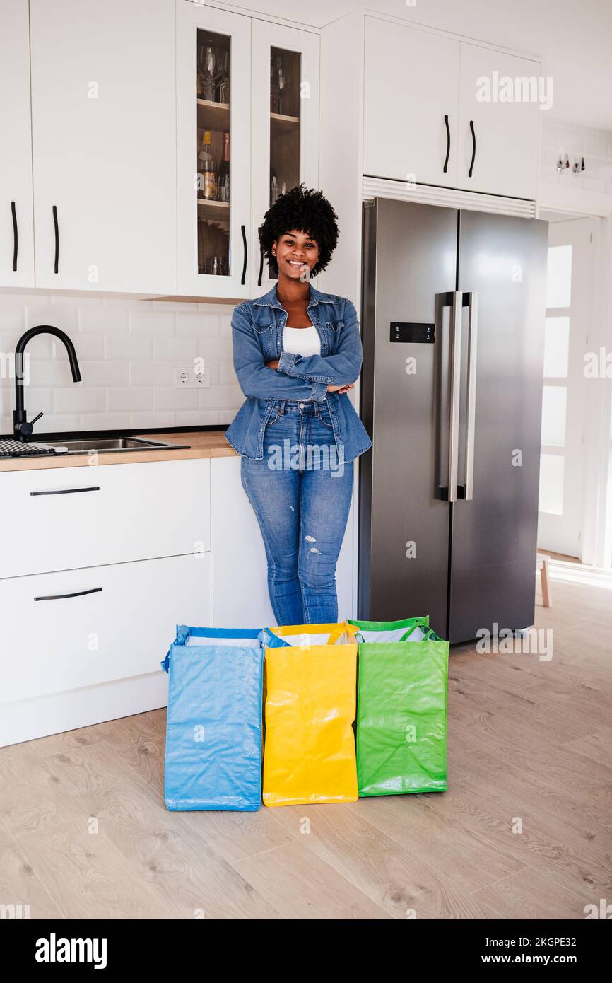 Donna felice con le braccia incrociate in piedi da borse riutilizzabili in cucina a casa Foto Stock