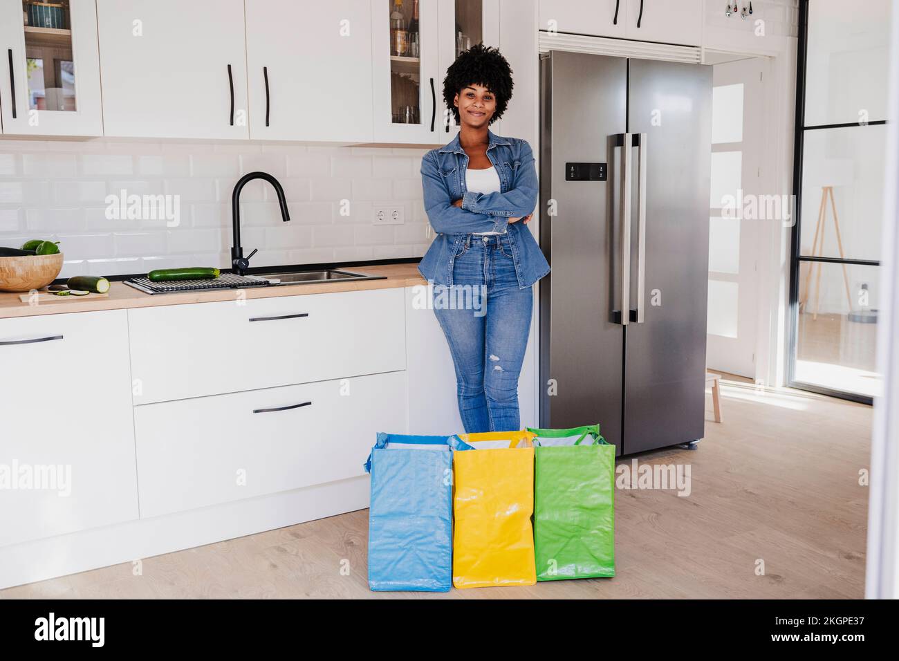 Donna sorridente con braccia incrociate in piedi da borse riutilizzabili in cucina a casa Foto Stock