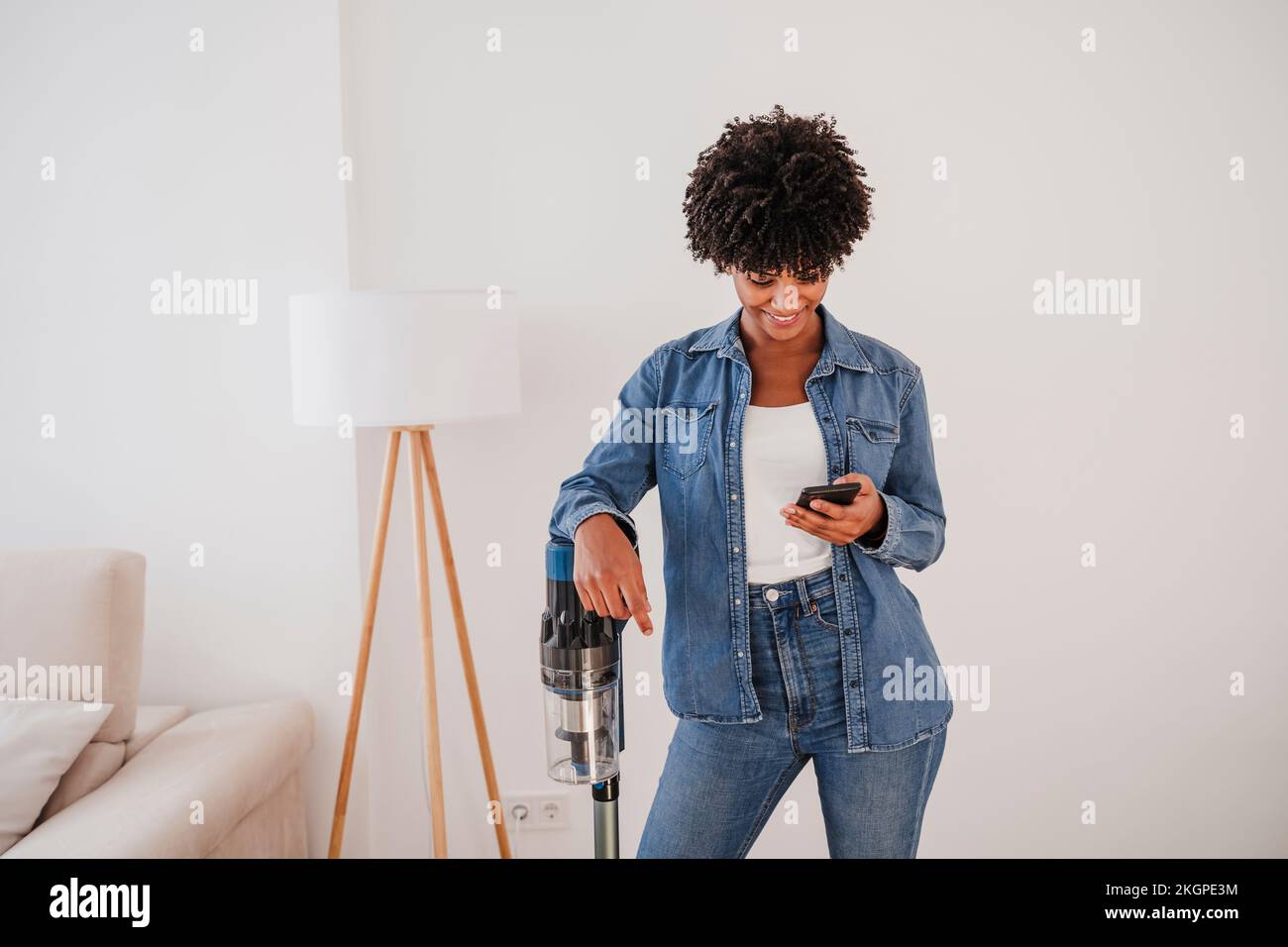 Donna sorridente in piedi con l'aspirapolvere e utilizzando lo smartphone a casa Foto Stock