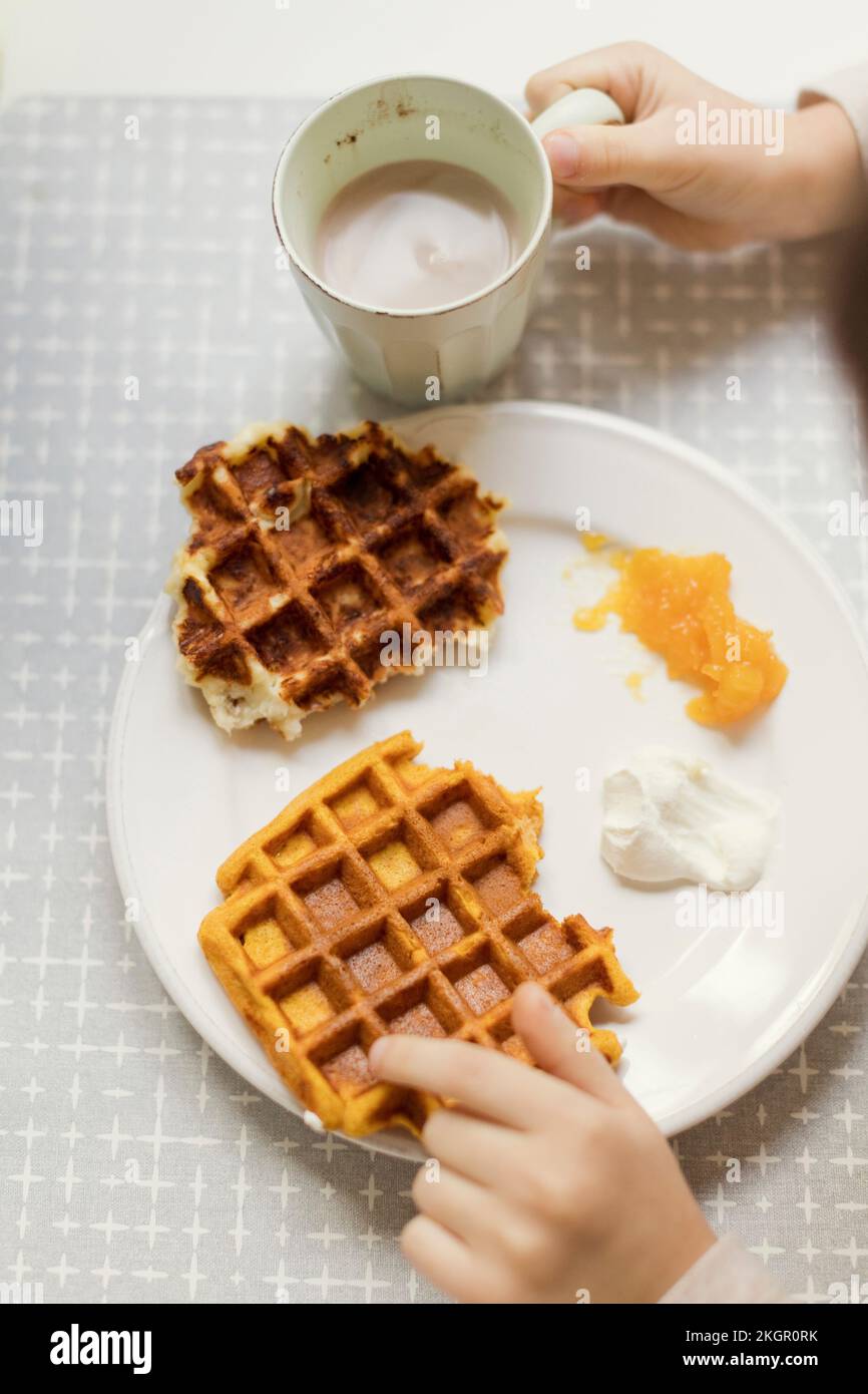 Mani di un ragazzo che tiene una tazza di cioccolata calda con waffle nel piatto del tavolo da pranzo Foto Stock