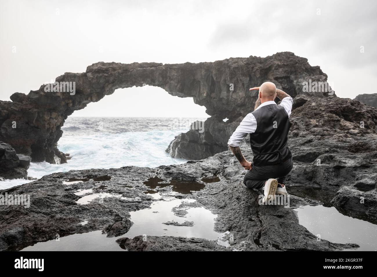 Uomo maturo che scherza gli occhi inginocchiati davanti all'arco di roccia Foto Stock