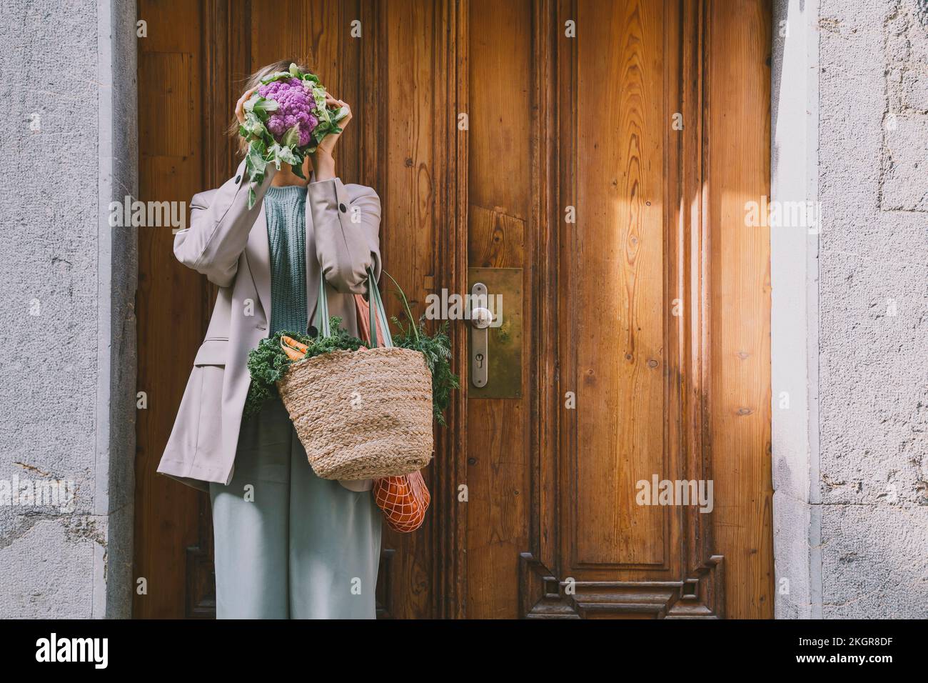 Donna che tiene borse coprire il viso con cavolfiore viola di fronte alla porta Foto Stock