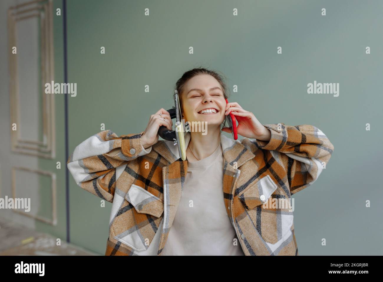Donna felice che tiene l'attrezzo di lavoro e lo smartphone sulle orecchie a casa Foto Stock
