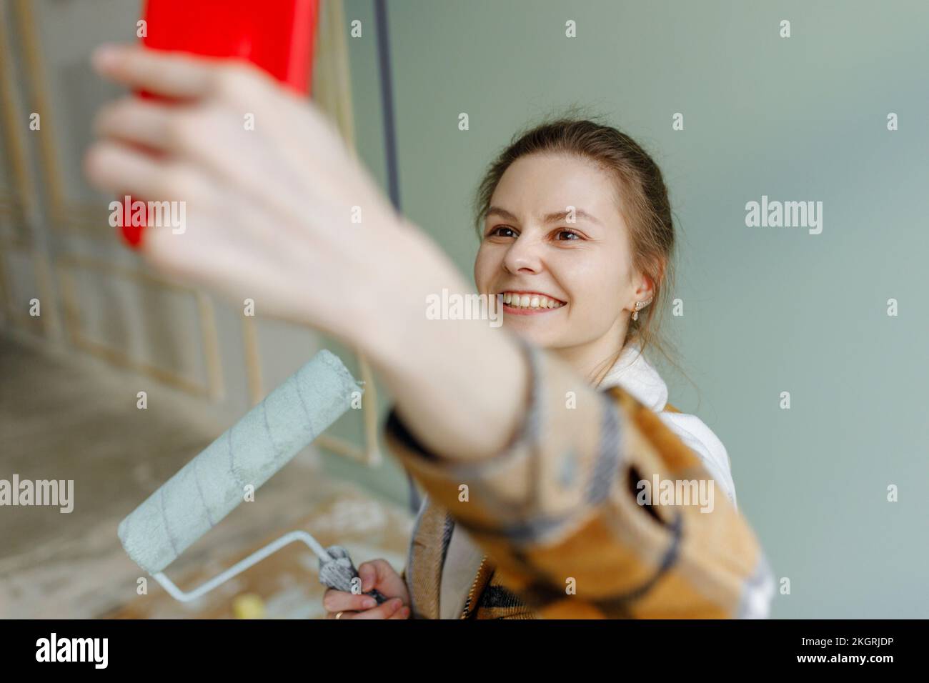 Donna sorridente con rullo di vernice che prende selfie sullo smartphone a casa Foto Stock