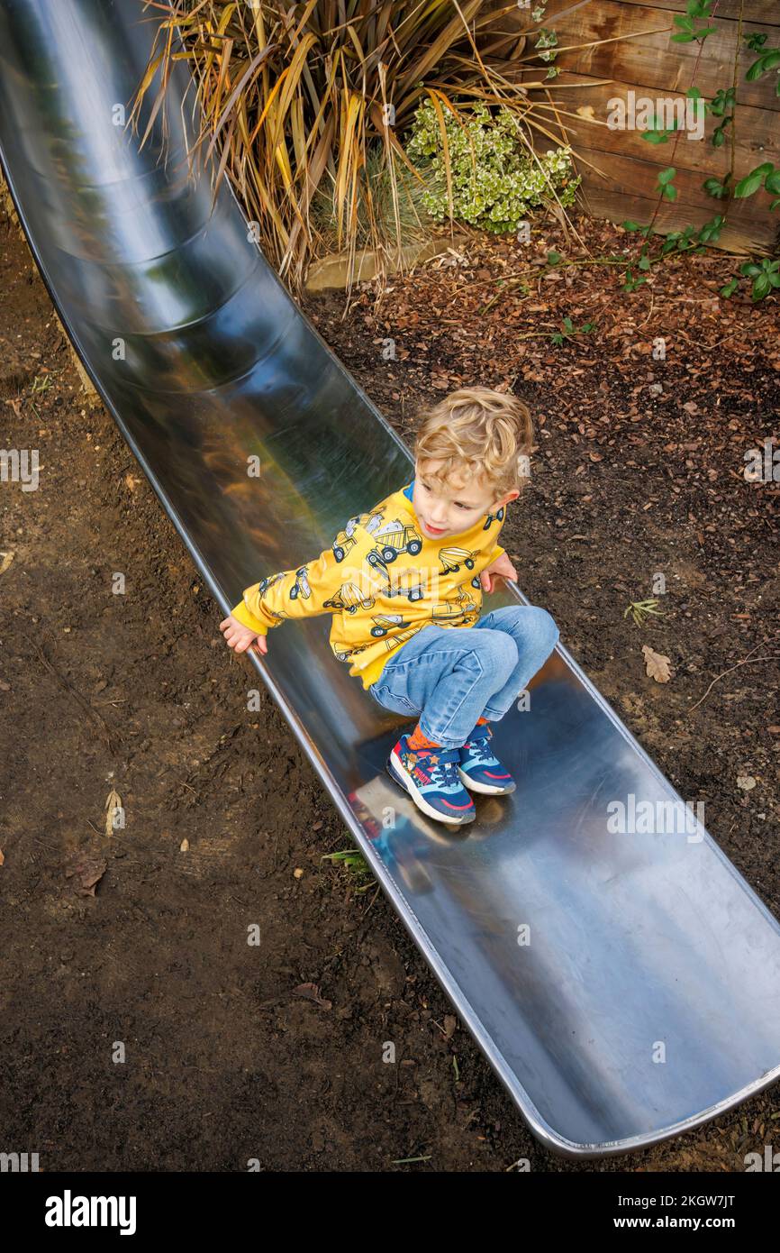 Un piccolo ragazzo caucasico biondo (di 4 anni) che indossa un top giallo e jeans blu in fondo a uno scivolo mentre gioca a Woking Park. Woking, Surrey Foto Stock