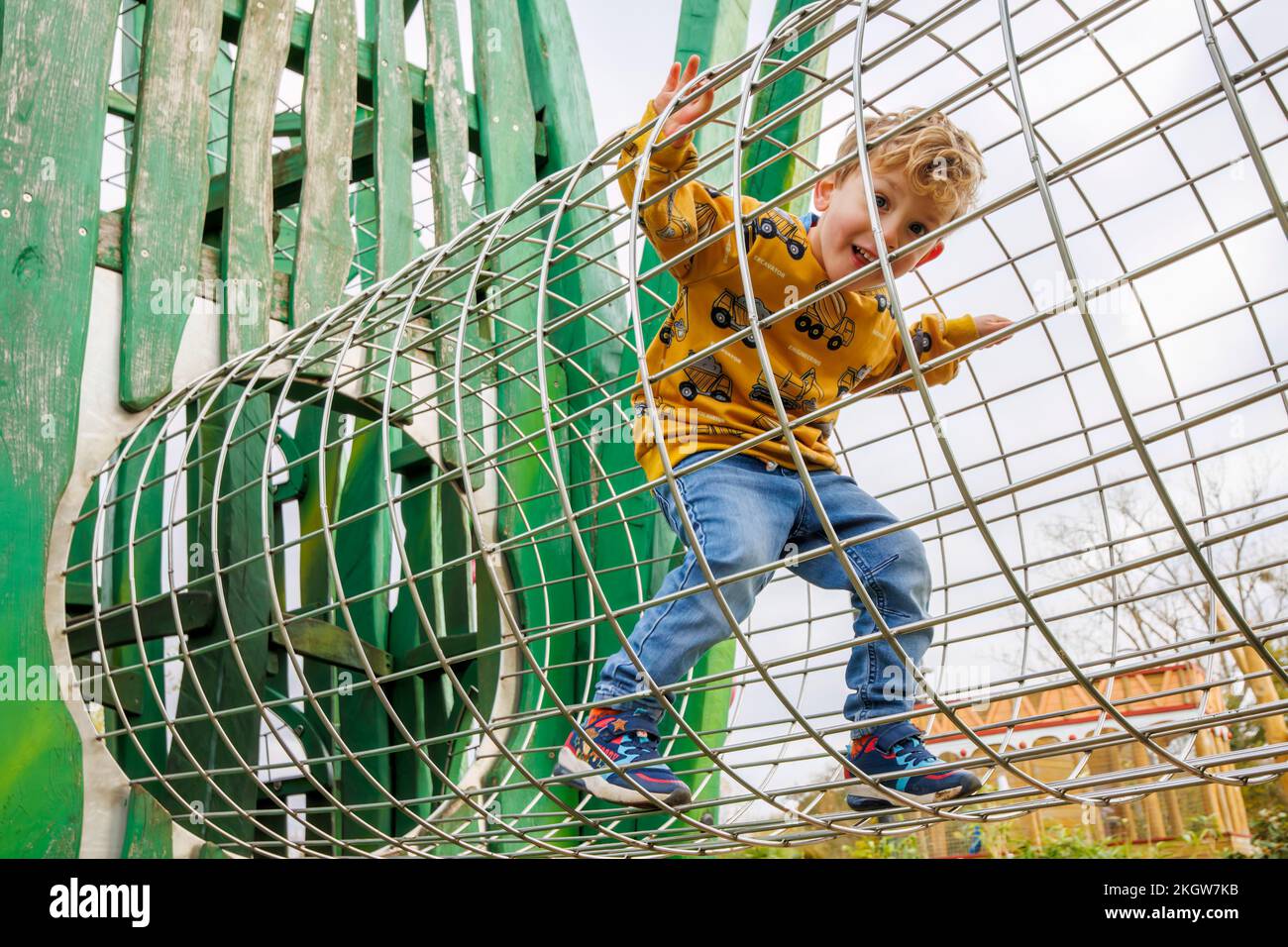 Un piccolo ragazzo caucasico biondo (di 4 anni) che indossa una parte superiore gialla e jeans blu cammina attraverso un cilindro di filo mentre gioca a Woking Park,. Woking, Surrey Foto Stock
