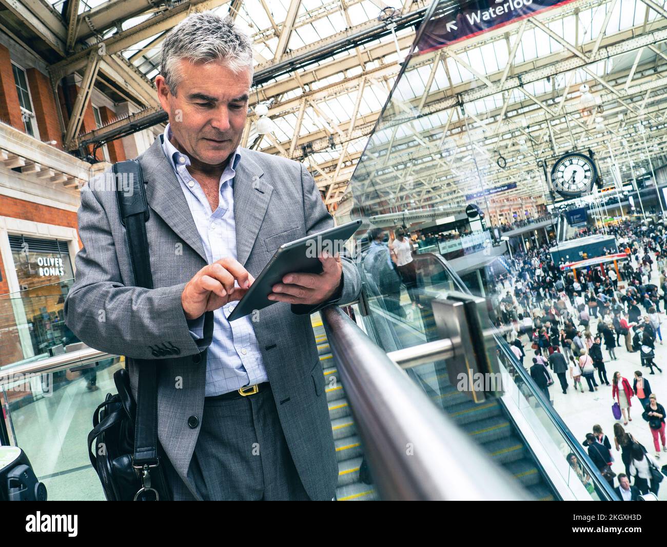 Uomo d'affari con iPad che utilizza un tablet intelligente su un'affollata stazione ferroviaria di Waterloo, atrio ferroviario con orologio, lavoro in movimento. Londra Regno Unito Foto Stock
