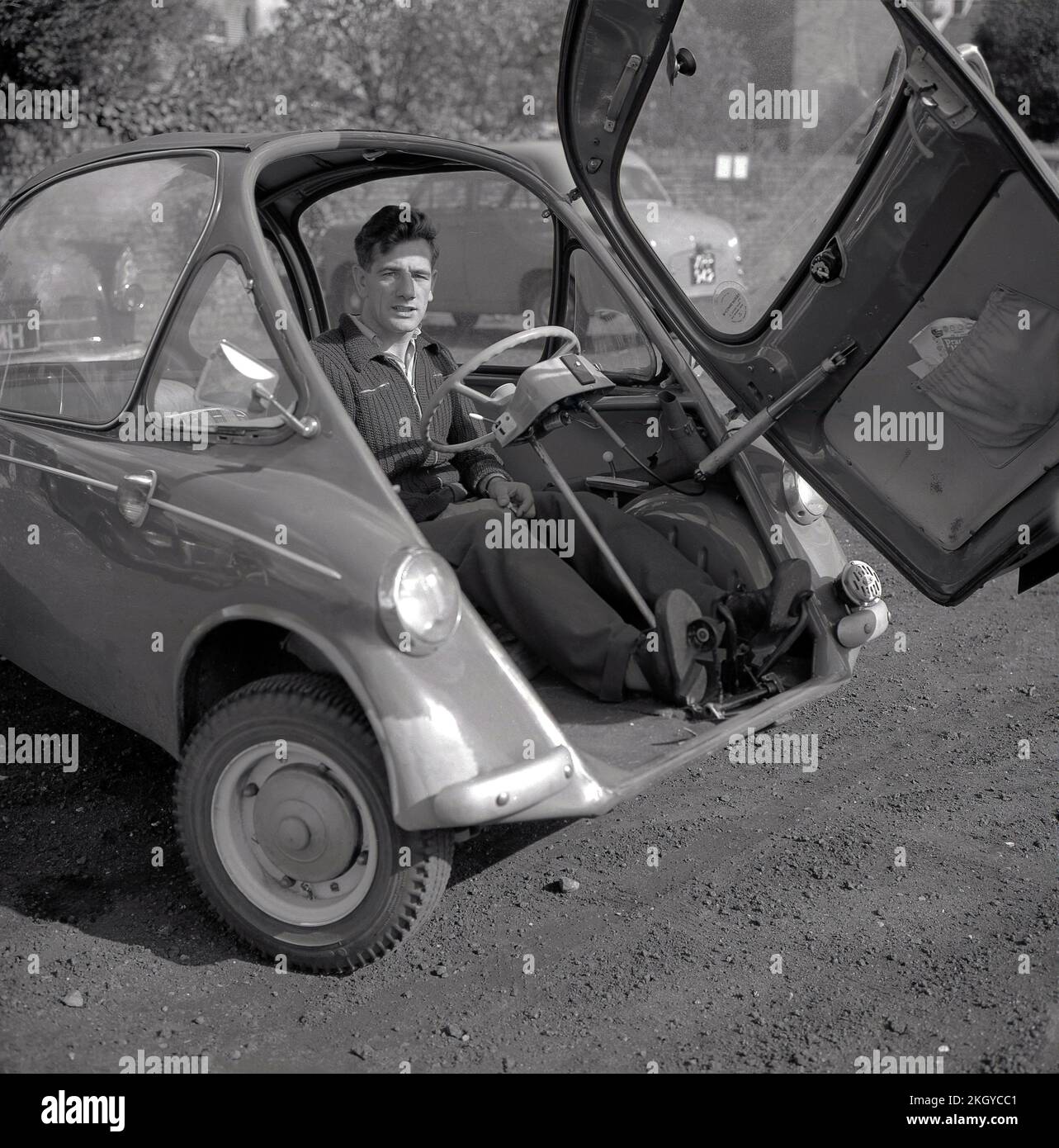 1960s, storico, fuori in un parcheggio di ghiaia, un uomo seduto all'interno di una piccola auto dell'epoca, una bolla auto, una BMW Isetta, che aveva una porta di apertura anteriore, Wycombe, Inghilterra, Regno Unito. Costruita per la prima volta da ISO Spa in Italia nel 1953, questa piccola auto è stata costruita su licenza in diversi paesi, tra cui BMW di Germania fino al 1962 e nota come la 'Bubble Car'. Foto Stock