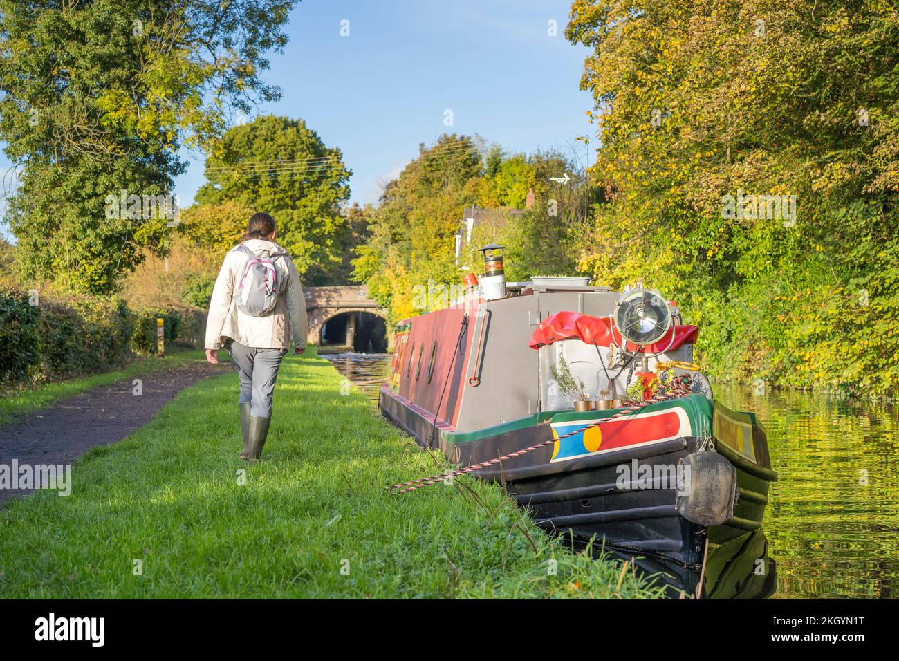 Vista posteriore di una donna che cammina lungo un canale con una barca ormeggiata in una soleggiata giornata autunnale. Foto Stock