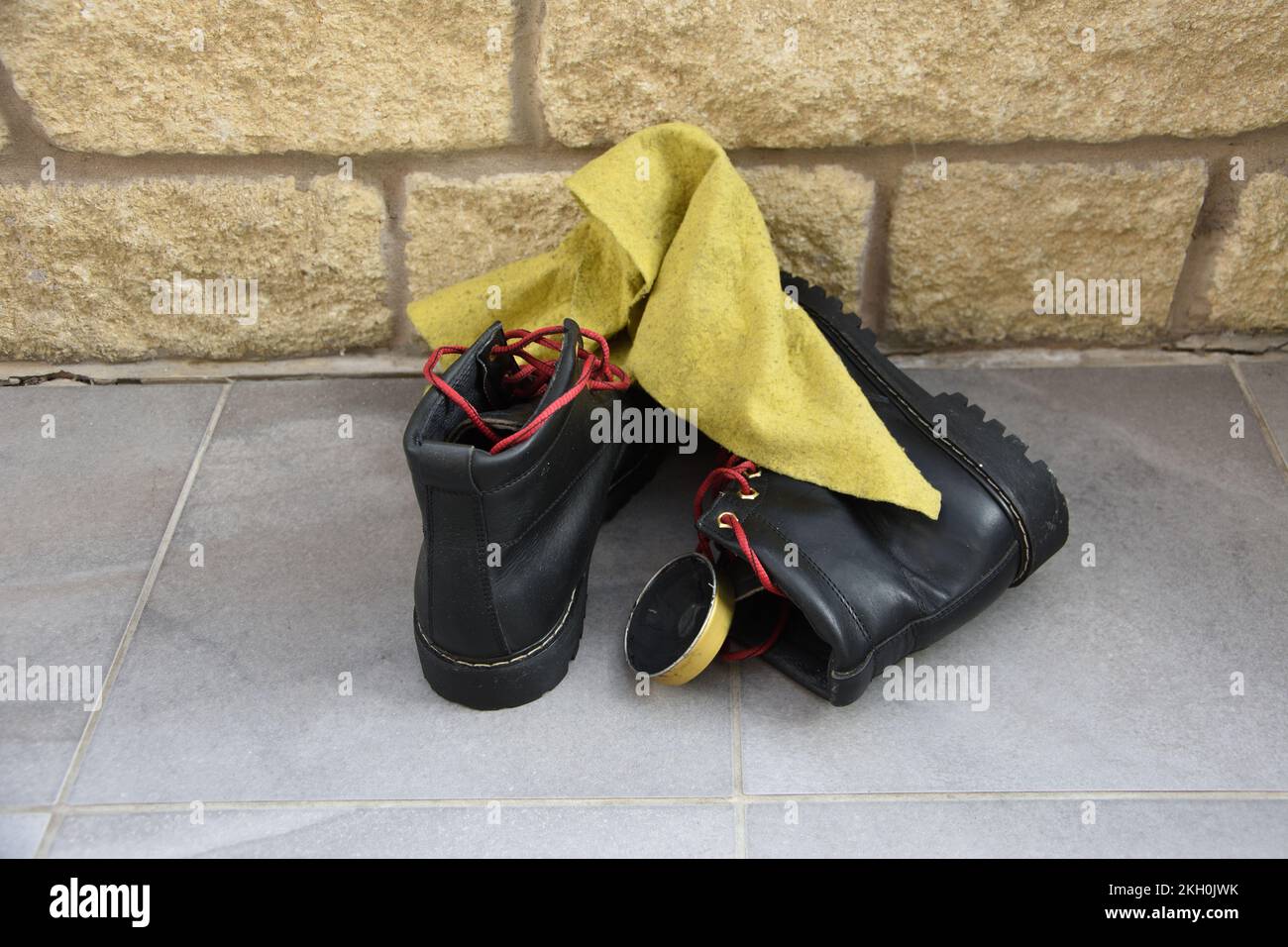 Stivali da passeggio neri con lacci rossi in attesa di pulizia su pavimento piastrellato con mattoni in pietra sul retro e un piumino giallo con una latta di lucidatura scarpe. Dopo Foto Stock