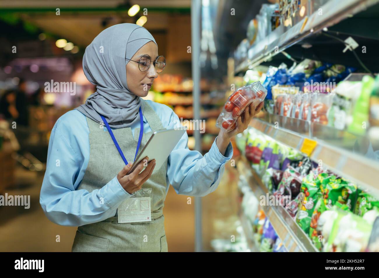 Una donna musulmana in un hijab, un operaio del supermercato controlla i prodotti sugli scaffali e in frigoriferi, una donna con un tablet e un grembiule usa un tablet per rivedere i prodotti. Foto Stock