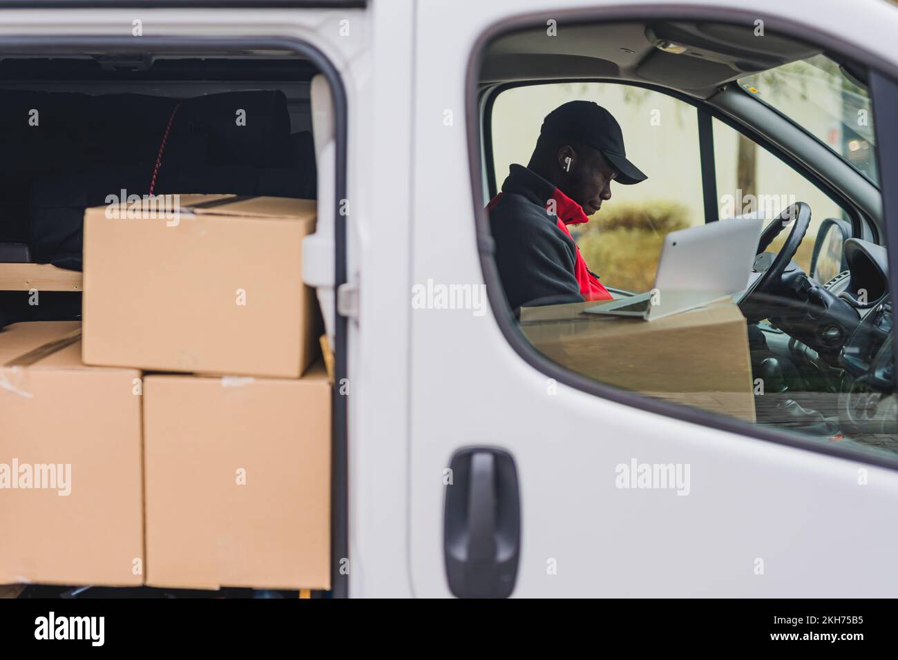 Corriere moderno. Concetto di consegna del pacchetto di shopping online. Furgone di consegna bianco con sportelli laterali aperti che rivelano molti pacchi non consegnati. Deliveryman nero adulto in cappello scuro e giacca rossa. Computer portatile Silver utilizzato per fornire gli indirizzi. . Foto di alta qualità Foto Stock