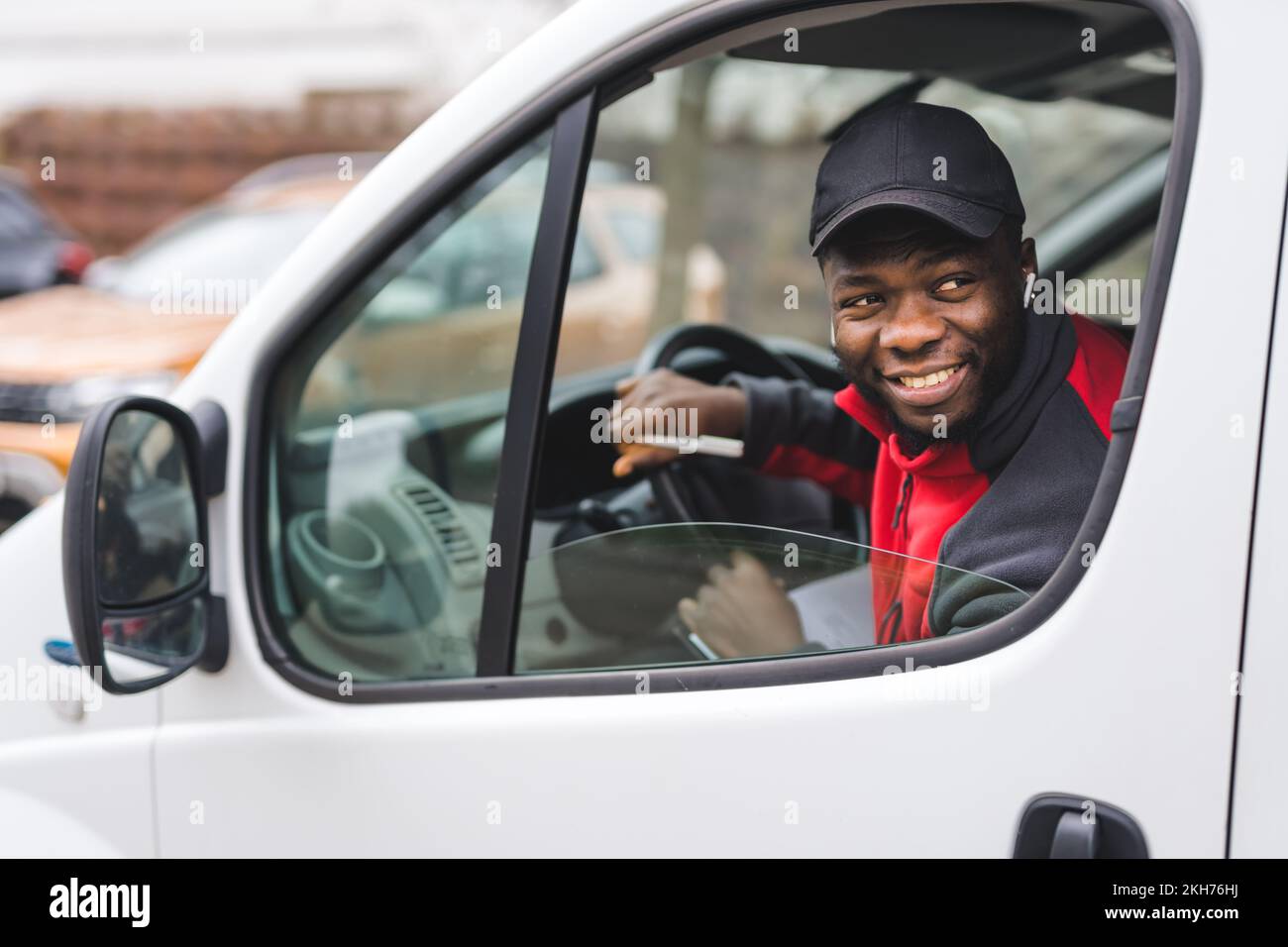 Giovane adulto nero addetto alla consegna seduto sul sedile del conducente di un furgone bianco che si inclina fuori dalla finestra sorridendo. Servizio di consegna. Scatto orizzontale. Foto di alta qualità Foto Stock