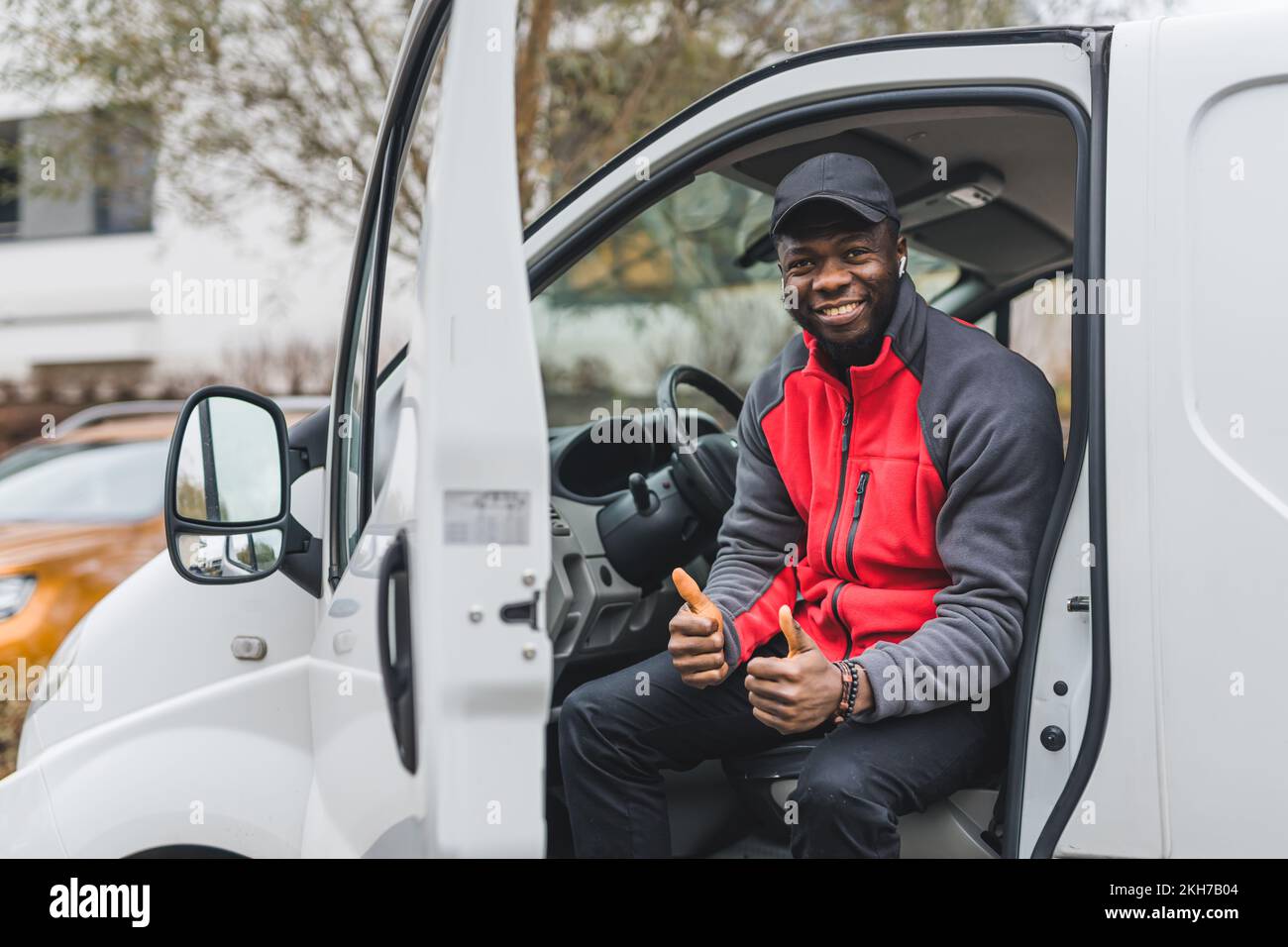 Giovane uomo di consegna nero adulto che indossa un pullover rosso e un cappuccio nero seduto nella porta del furgone bianco sorridente mostrando i pollici fino alla fotocamera. Scatto orizzontale all'aperto. Foto di alta qualità Foto Stock