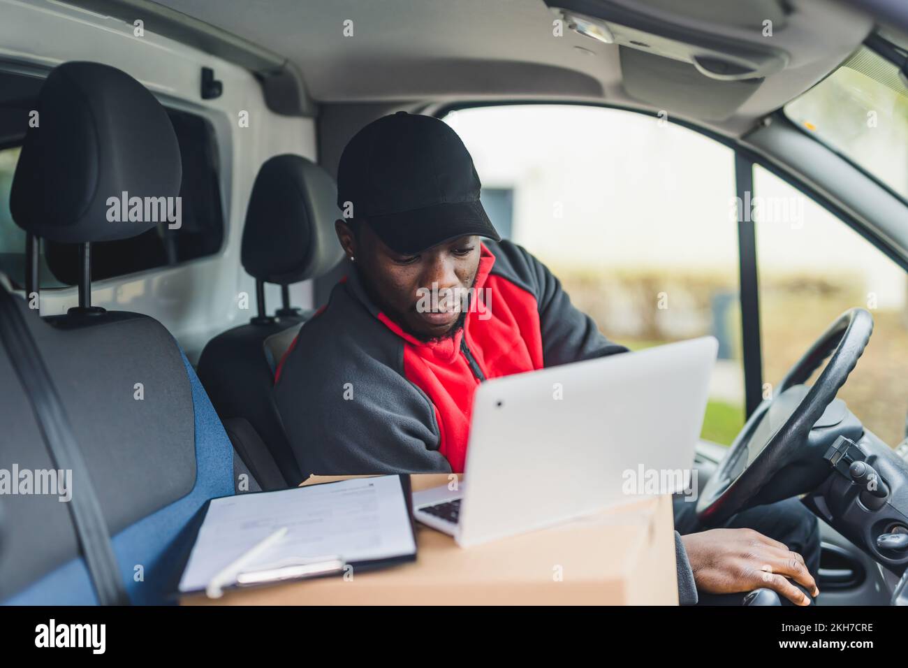 Vista a bordo dell'auto dell'uomo nero occupato e focalizzato di mezza età in uniforme che utilizza il suo laptop argentato per superare i pacchi successivi. L'uso del veicolo - furgone bianco. Foto di alta qualità Foto Stock