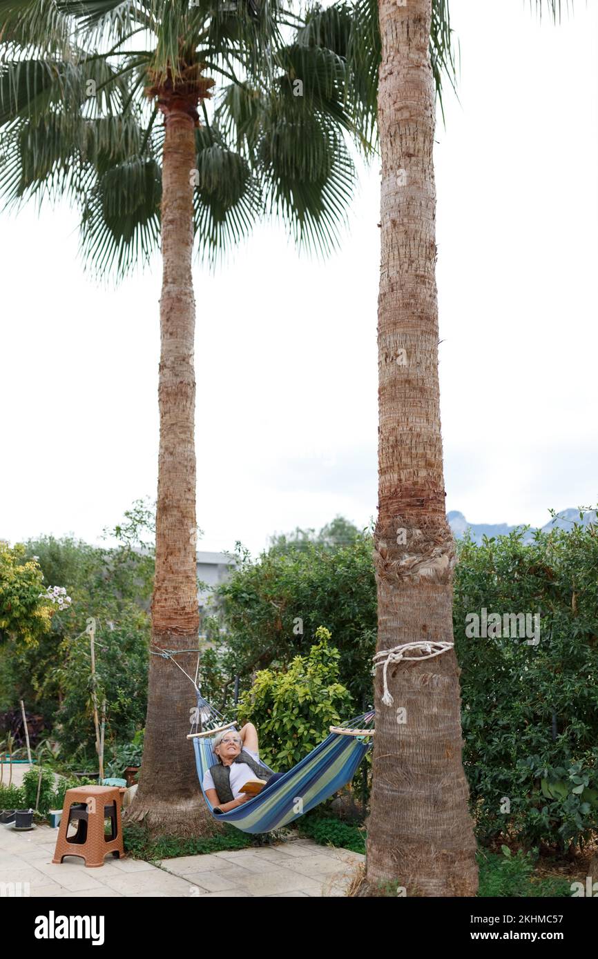 Donna sorridente di capelli di buon aspetto grigio indossare occhiali mentre la lettura in amaca nel giardino estivo sotto le palme Foto Stock