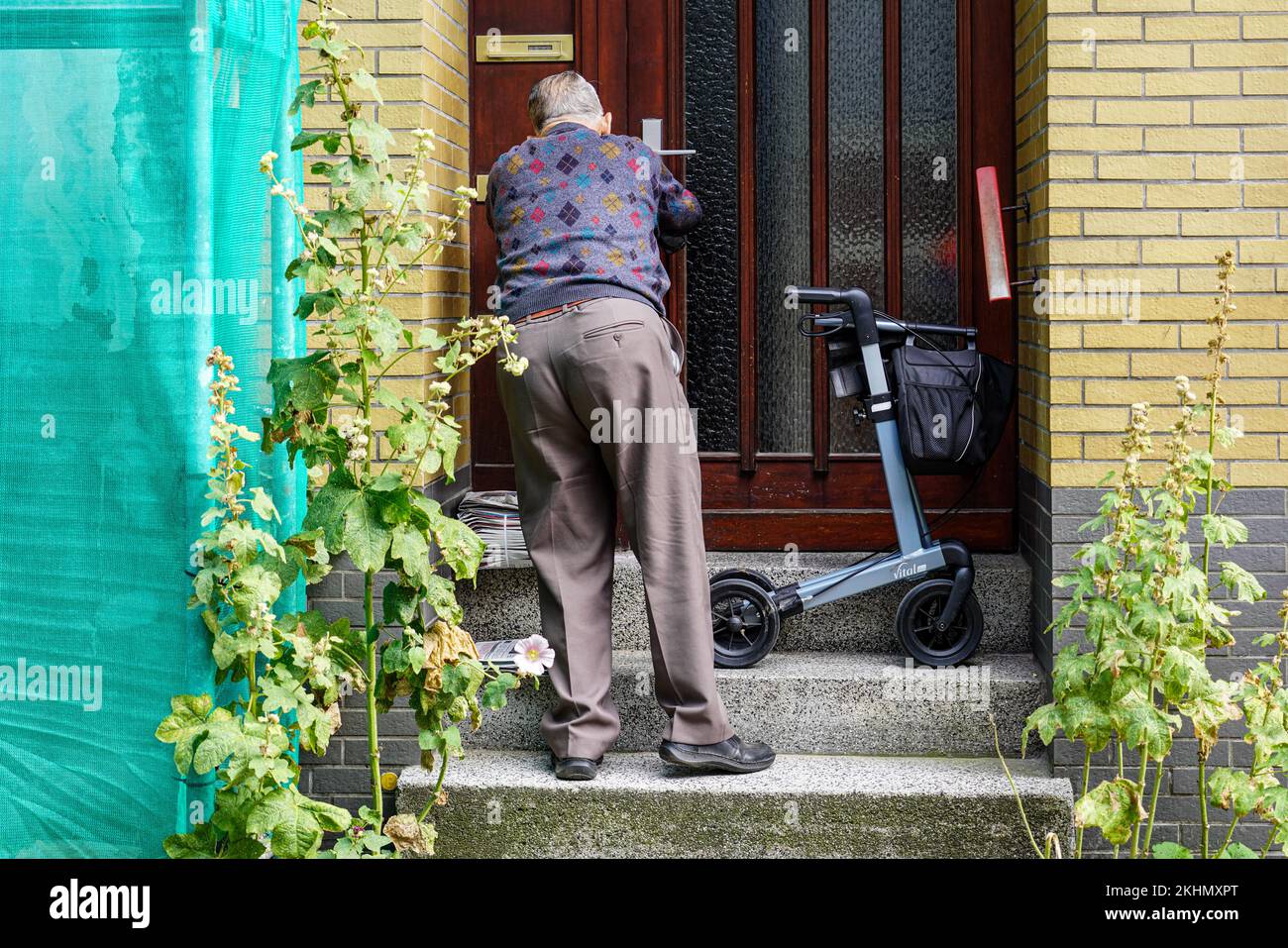 Un Signore anziano con una disabilità ambulante deve andare su tre punti per sbloccare la porta del suo appartamento. Ha già portato il suo camminatore su i gradini. Foto Stock