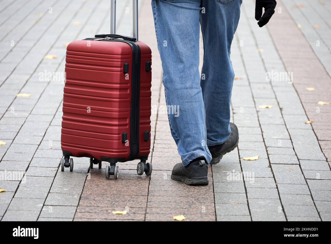 Valigia rossa su ruote e gambe maschio sul marciapiede. Uomo con bagagli che cammina lungo la strada autunnale, concetto di viaggio Foto Stock