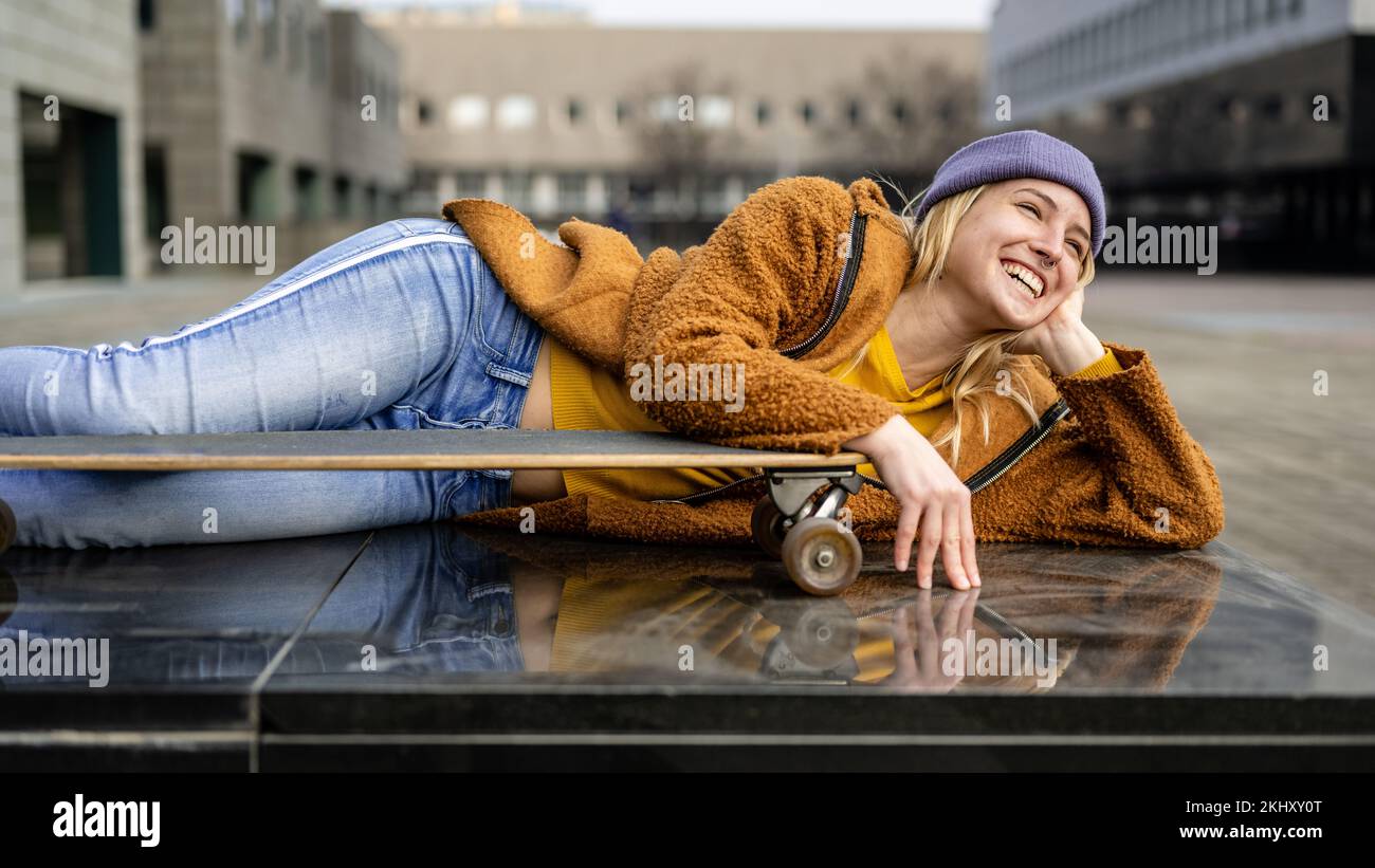 Lungo skater femmina di bordo che ha un riposo su un muro in contesto urbano, felice toothy sorridente ragazza portrai, generazione z Foto Stock