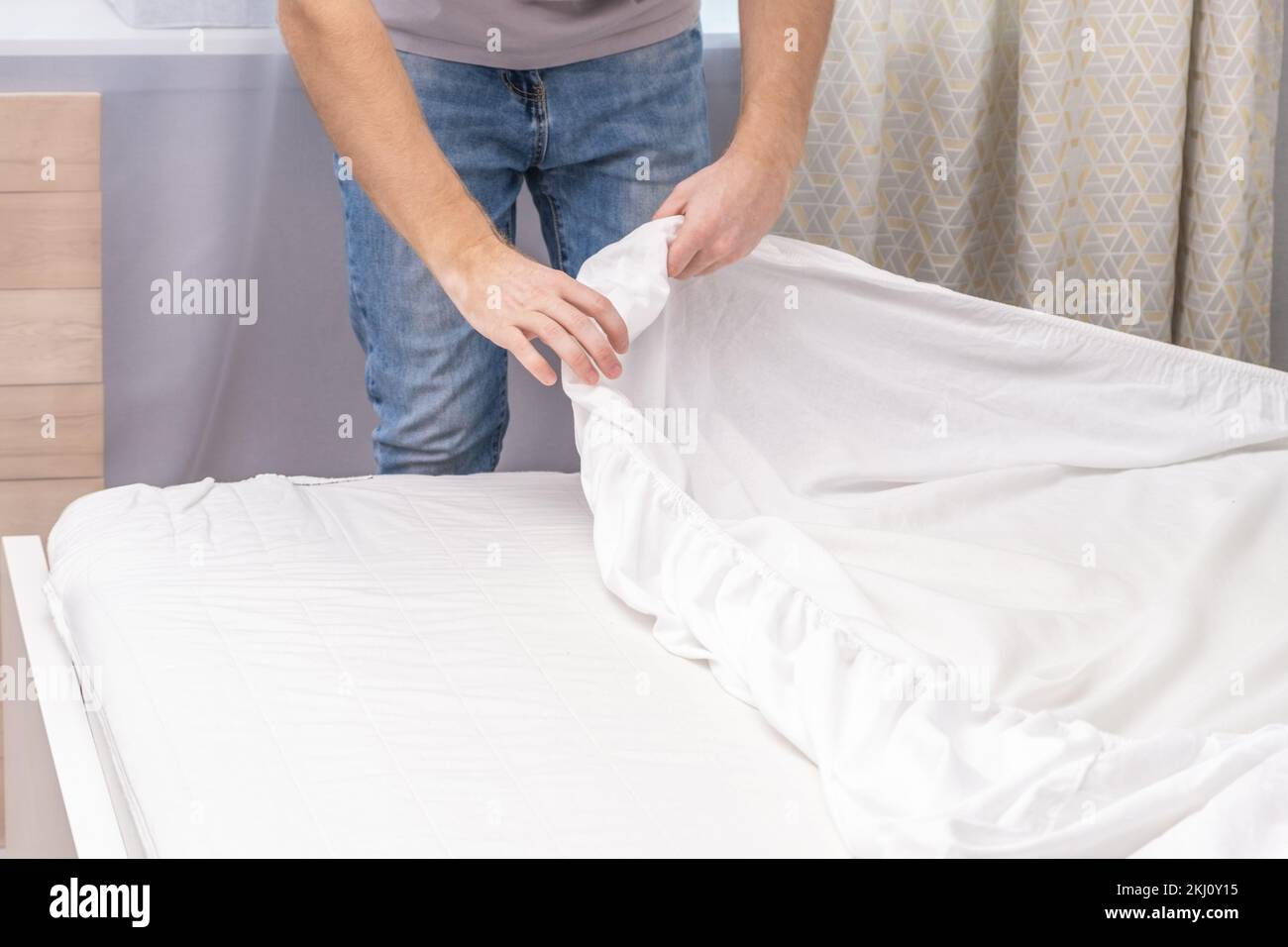 Un uomo fa il suo letto accogliente con fresche lenzuola bianche e luminose. Fare il letto con lenzuola fresche da un uomo bianco. Il giorno del cambio della biancheria. Il giorno del lavaggio biancheria nella lavanderia. Foto Stock
