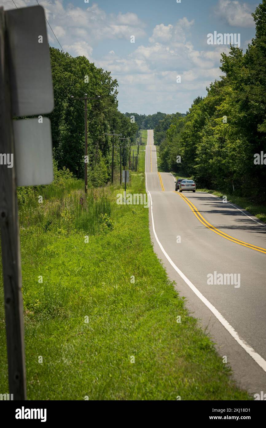 Lunga strada asfaltata con linee gialle e bianche in America, attraversa una piccola valle circondata da alberi ed erba Foto Stock