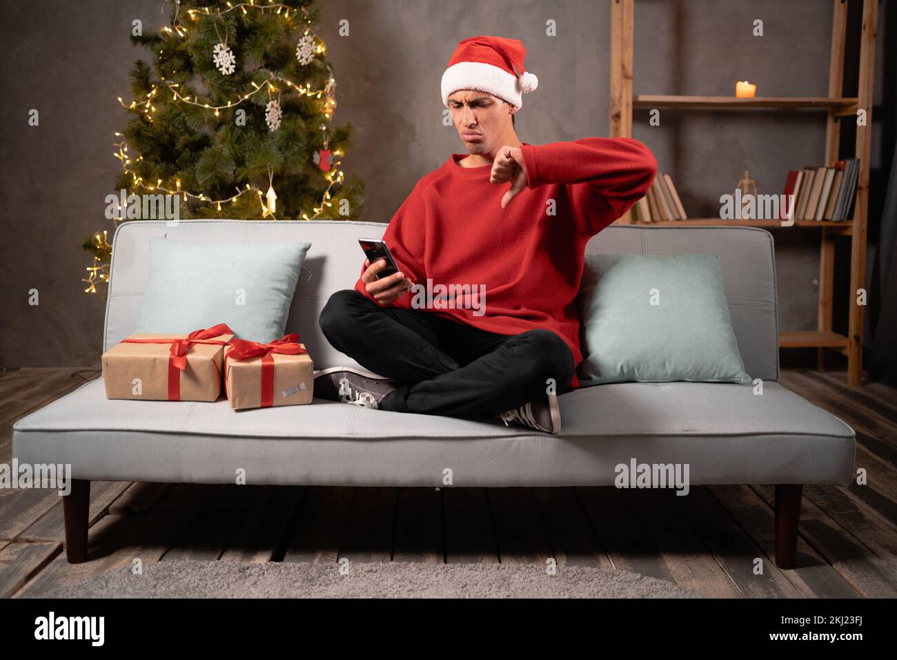 Un giovane insoddisfatto a casa di santa Hat sul divano che utilizza l'app per smartphone per consegnare le merci mostra un gesto verso il basso. Molto povero regalo di Natale Deli Foto Stock
