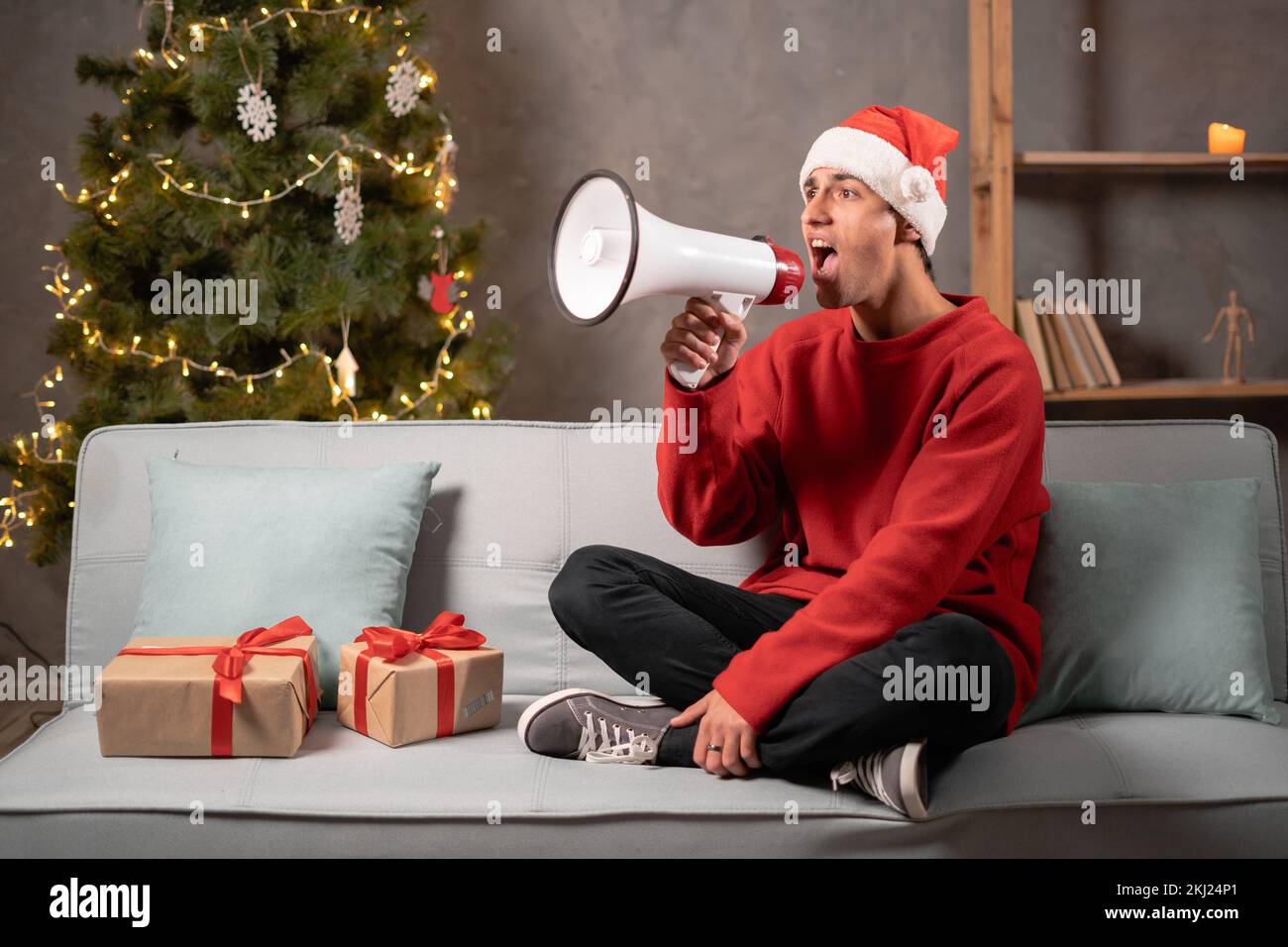 Giovane uomo felice nel cappello di Babbo Natale che urla usando il megafono mentre si siede a casa. Condividere notizie sulle vendite stagionali. Concetto di vendita di Natale Foto Stock