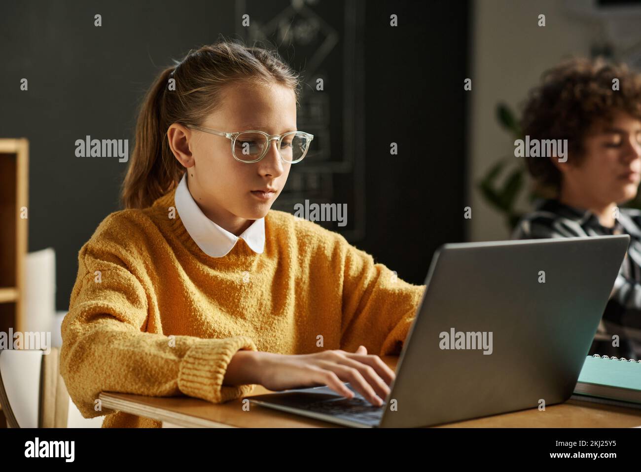 Studentessa di occhiali che si concentra sul suo lavoro online sul computer portatile mentre si siede alla scrivania a lezione a scuola Foto Stock