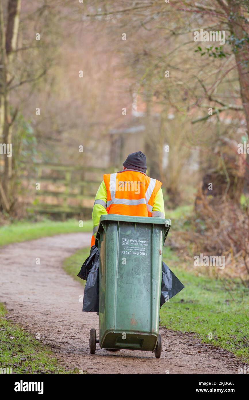 Vista posteriore del lavoratore del consiglio maschile in giacca ad alta visibilità e cappello che tira il cestino verde di riciclaggio dietro di lui nel parco pubblico. Bin MAN Regno Unito. Foto Stock