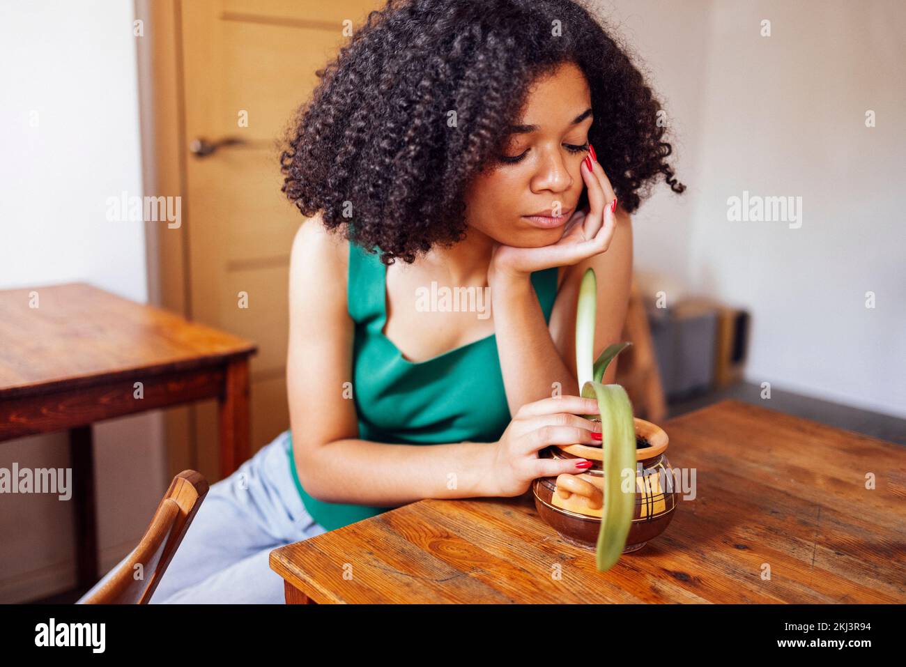Giovane donna triste di etnia afroamericana indossa la t-shirt verde tiene l'albero della pianta della casa seduto sul soggiorno Foto Stock