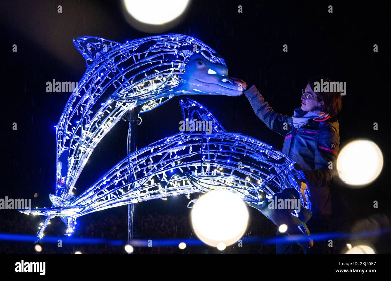 24 novembre 2022, Sassonia, Lichtenwalde: Una donna guarda due delfini illuminati nel giardino barocco del Palazzo e del Parco di Lichtenwalde durante un'anteprima dello spettacolo LUMAGICA. In un tour di circa 1,5 km, i visitatori possono sperimentare un mondo di luce invernale con più di 300 oggetti illuminati, produzioni di luce sincronizzate con la musica e elementi interattivi Il tema della messa in scena si basa sulla leggenda del cavaliere Harras, che sta cercando il suo cavallo. I visitatori camminano attraverso una foresta incantata con piante luminose e insetti nel parco del castello, entrano in un regno sottomarino segreto della Zschopa Foto Stock