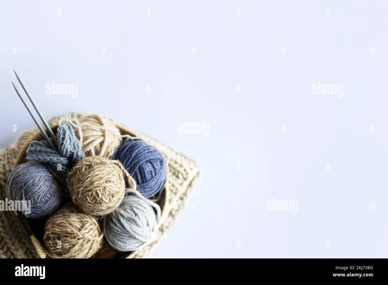 Palle grigie, marroni e blu di fili da maglieria in un cesto beige con aghi da maglieria e una pila di cappelli da maglieria per inverno e autunno, piano cottura da maglieria Foto Stock