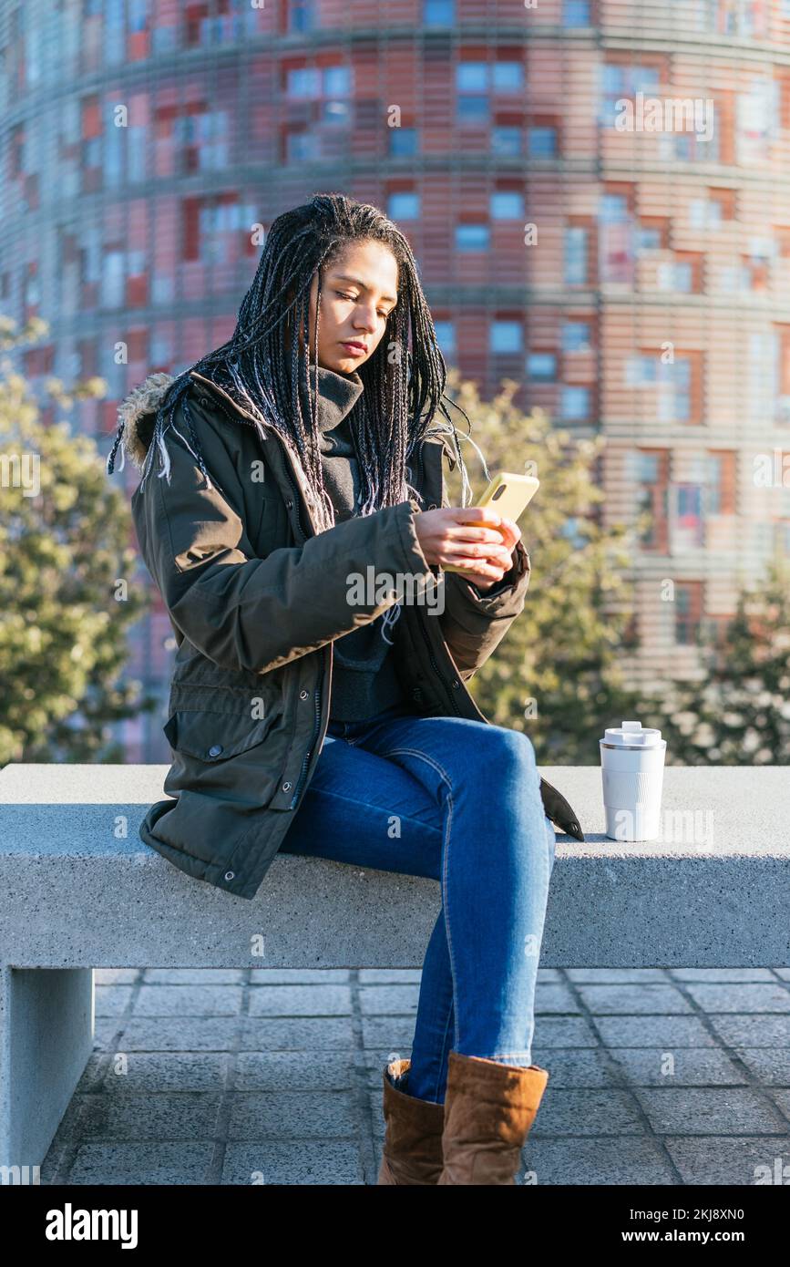 Donna ispanica focalizzata in outerwear messaggi di testo sul cellulare mentre si siede su una panca di pietra con thermos di caffè caldo in città Foto Stock